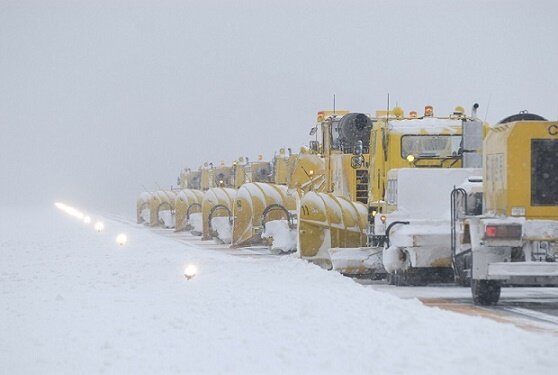 降雪時の空港滑走路で、複数の空港用除雪車両が隊列を組んで除雪作業を行っている様子。視程が低下する中、滑走路灯を確保しながら作業している。